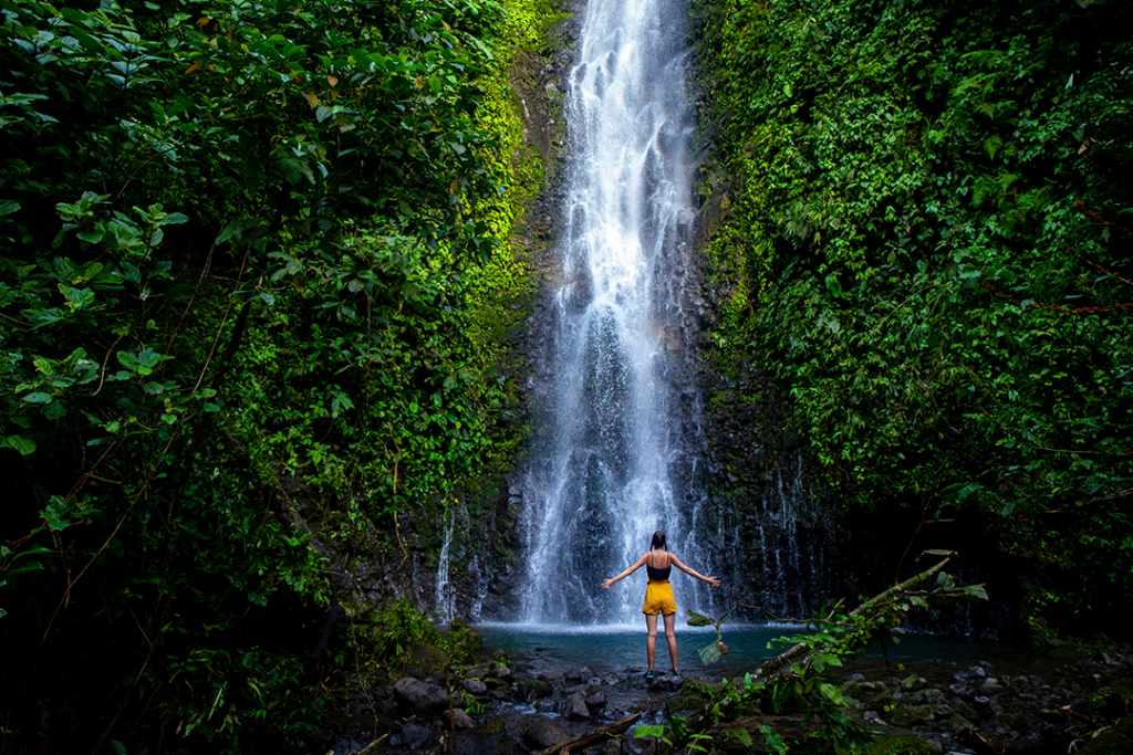 brave girl stands in front of a mighty waterfall with her hands raised in the air; celebration of a successful climb over a waterfall; tropical waterfall in Costa Rica; hidden gems of costa rica